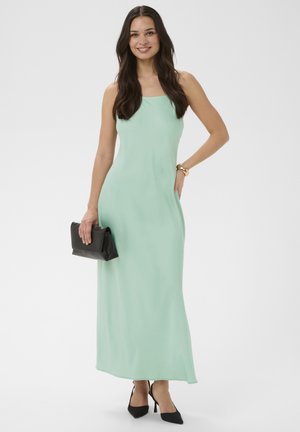 Femme souriante, portant une robe longue sans manches vert menthe, des talons pointus noirs, un bracelet en or, et tenant une pochette noire.