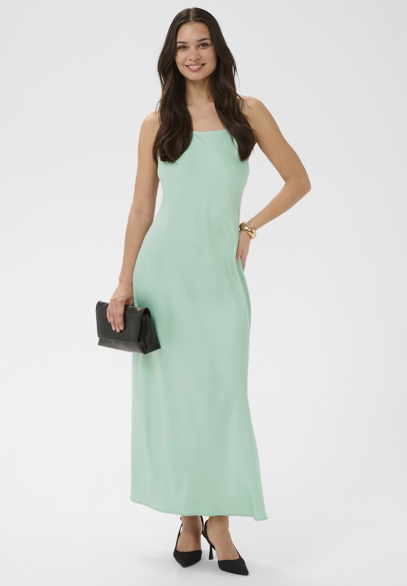 Femme souriante, portant une robe longue sans manches vert menthe, des talons pointus noirs, un bracelet en or, et tenant une pochette noire.