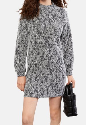 Femme portant une robe à manches longues noire et blanche à motifs, tenant un sac à main noir matelassé dans sa main droite.
