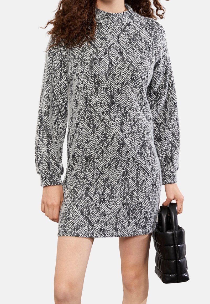 Femme portant une robe à manches longues noire et blanche à motifs, tenant un sac à main noir matelassé dans sa main droite.