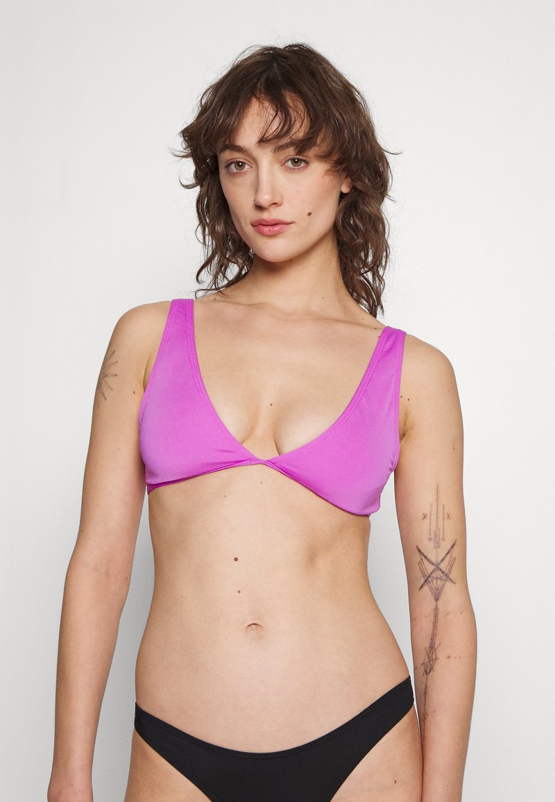 Donna con capelli castani mossi che indossa un reggiseno bikini rosa brillante e uno slip bikini nero, in piedi contro uno sfondo bianco uniforme.