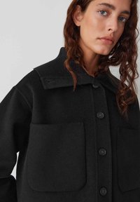 Veste en mélange de laine noire avec un col texturé, deux poches avant et des boutons proéminents sur le devant.