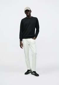 Μαύρο φούτερ, λευκό παντελόνι και μαύρα αθλητικά παπούτσια με λευκές ρίγες. Το καπέλο είναι μπεζ. Το μοντέλο στέκεται μπροστά σε ανοιχτό φόντο.