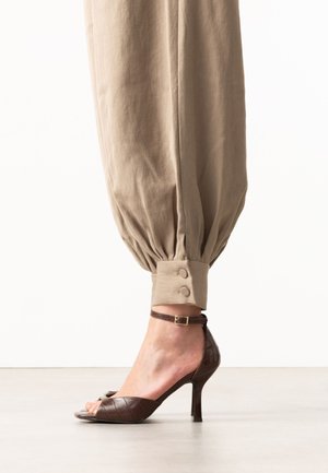 Beige pants with buttoned cuffs paired with brown high-heeled open-toe sandals with ankle straps on a light floor background.