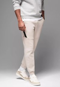 Svetle béžové tepláky s hladkou texturou, s černou akcentní kapsou a užším střihem, doplněné bílými a béžovými teniskami.