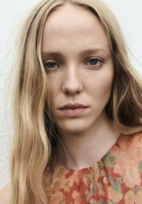 Portret osoby z długimi, falującymi blond włosami, ubraną w pomarańczową sukienkę w kwiatowy wzór z zielonymi akcentami, na jasnym tle.