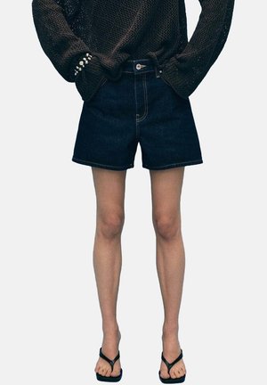Person iført sort strikket sweater, mørkeblå denimshorts og sorte flip-flops står med hænderne i lommerne.