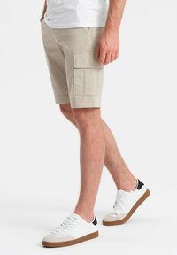 Shorts cargo beiges avec deux grandes poches latérales, devant plat, et ourlet se terminant au-dessus du genou. Associés à des baskets blanches avec un accent noir.