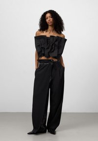 Zwarte off-shoulder top met gerimpelde textuur, gecombineerd met zwarte wijde broek. De broek heeft een geruite detail aan de zijkant en een gestrikte tailleband.