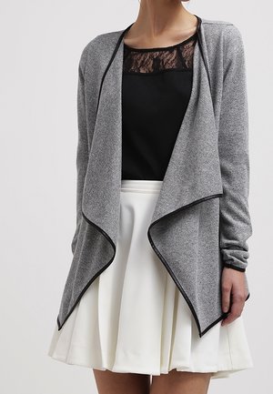 Kvinde iført en grå cardigan med åben front og sort kant over en sort blonde top og et hvidt plisseret skørt.