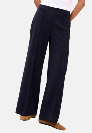 Isik, kes kannab kõrge vöökohaga, laiade säärega tumesiniseid pükse, mille esiküljel on õmblused, kombineeritud beežide madalate jalanõudega ja valge topiga, seisab valge tausta ees.