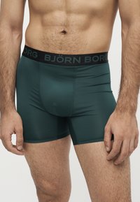 Homme portant un boxer ajusté vert foncé avec une ceinture noire portant l'étiquette Björn Borg, montré de la taille au milieu de la cuisse.
