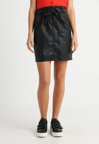 Jupe en simili cuir noire avec taille cintrée et détail en nœud. Présente une coupe droite et une texture lisse, associée à des baskets à semelle blanche.