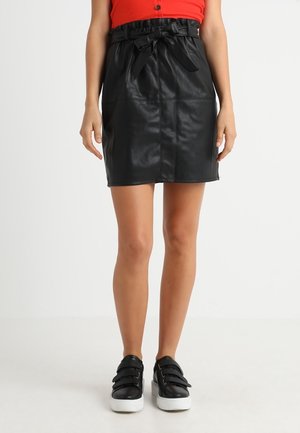 Jupe en simili cuir noire avec taille cintrée et détail en nœud. Présente une coupe droite et une texture lisse, associée à des baskets à semelle blanche.