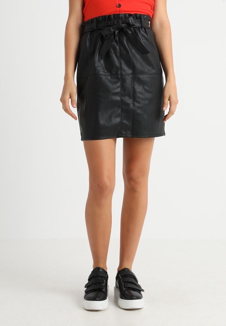 Jupe en simili cuir noire avec taille cintrée et détail en nœud. Présente une coupe droite et une texture lisse, associée à des baskets à semelle blanche.