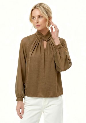 Mujer rubia con blusa de manga larga verde oliva con escote fruncido en forma de ojo de cerradura y pantalones blancos, tocando un pendiente de aro dorado.