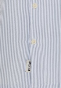 Jasnoniebieska koszula w białe paski wykonana z materiału, z okrągłymi guzikami i metką z napisem "HOLLISTER" na dole.