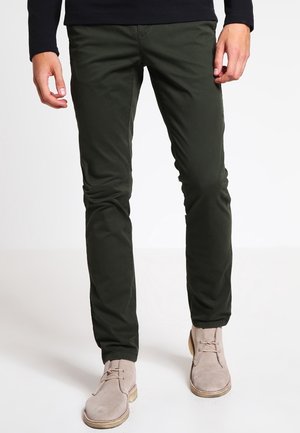 Mann trägt dunkelgrüne, eng anliegende Hose und beige Wildleder-Schnürstiefeletten, steht vor weißem Hintergrund.