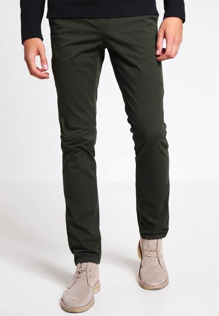 Homme portant un pantalon vert foncé ajusté et des bottines beiges en daim à lacets, debout sur un fond blanc.