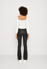 Frau mit lockigem Haar, die ein weißes Top mit quadratischem Ausschnitt, schwarze enganliegende Schlaghosen und weiße Schuhe mit dicker Sohle trägt, und von hinten schaut.