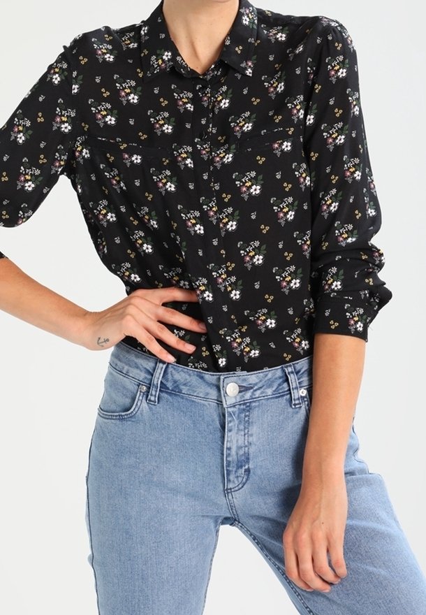 Femme portant une chemise noire à fleurs à manches longues rentrée dans un jean bleu clair taille haute, posant avec une main sur la hanche.