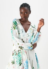 Abito bianco con motivi floreali in verde, blu e rosa. Presenta un profondo scollo a V, maniche lunghe e dettagli in pizzo lungo le cuciture.