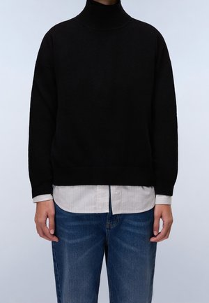 Personne portant un pull à col roulé noir superposé à une chemise blanche rayée sortie et un jean bleu, debout devant un fond clair.
