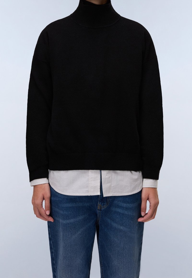 Personne portant un pull à col roulé noir superposé à une chemise blanche rayée sortie et un jean bleu, debout devant un fond clair.
