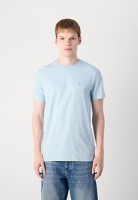 Helles blaues T-Shirt mit kurzen Ärmeln aus weicher Baumwolle, Rundhalsausschnitt und kleinem Logo auf der linken Brust; kombiniert mit ausgebleichtem blauen Jeans.