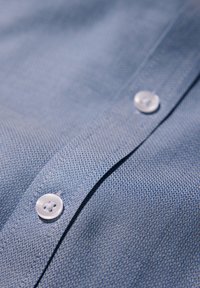 Vaaleansininen nappikaulusmainen paita, jossa on tekstuuri ja kudottu kuvio. Kaksi pyöreää, läpinäkyvää nappia näkyy napituksessa.