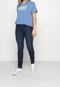 Kvinna i blå Levi's t-shirt, mörka tajta jeans och vita platåskor som står mot en ljus, enfärgad bakgrund.