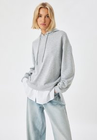 Sudadera gris con capucha, hecha de una tela suave y texturizada. Lleva puesta sobre una camisa blanca, combinada con unos jeans de pierna ancha de color azul claro.