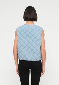 Αμάνικο τζιν γιλέκο σε ανοιχτό μπλε χρώμα με διακοσμητικό σχέδιο ρόμβου. Διαθέτει στρογγυλή γραμμή λαιμού και κατεβασμένο μήκος. Φορώντας με μαύρο παντελόνι.