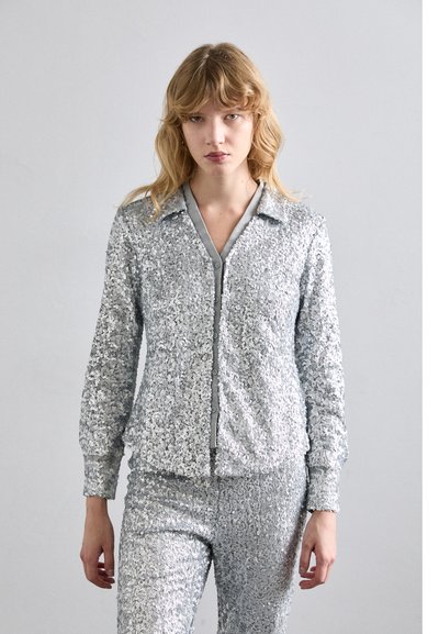 Chándal de lentejuelas plateadas con chaqueta con cuello, cremallera frontal y pantalones a juego. Textura suave con acentos brillantes y reflectantes.