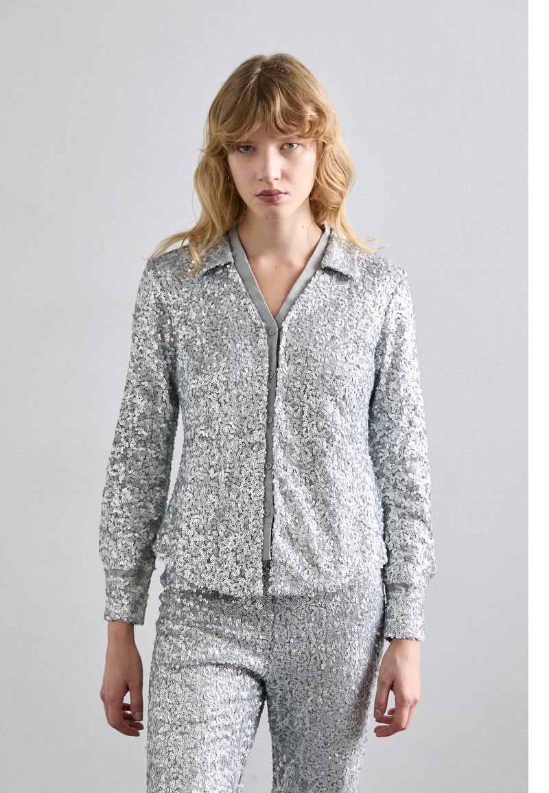 Hõbedane sädelev tracksuit, mis sisaldab kraega jakki, esimest lukku ja sobivaid pükse. Sile tekstuur läikivate ja peegeldavate aktsentidega.