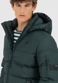 Giacca a vento verde scuro con finitura texture, chiusura con zip, cappuccio regolabile e una patch sulla manica. Presenta una maglietta a righe sotto.