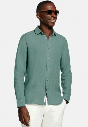 Lächelnder Mann mit dunkler Sonnenbrille, einem türkisfarbenen, langärmeligen Hemd mit Knöpfen und weißer Hose, die Hände in den Taschen, vor einem schlichten Hintergrund.