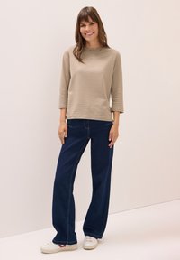 Helles beige gestreiftes Langarmoberteil, kombiniert mit weitem, dunkelblauen Jeans. Das Model trägt weiße Sneaker mit roten Akzenten. Einfaches, lässiges Outfit.