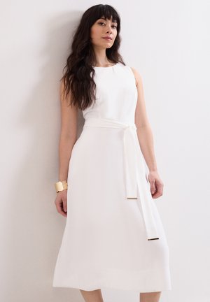 Woman with long dark hair wearing a sleeveless white midi dress with a tied belt and gold cuff bracelet standing against a plain wall.