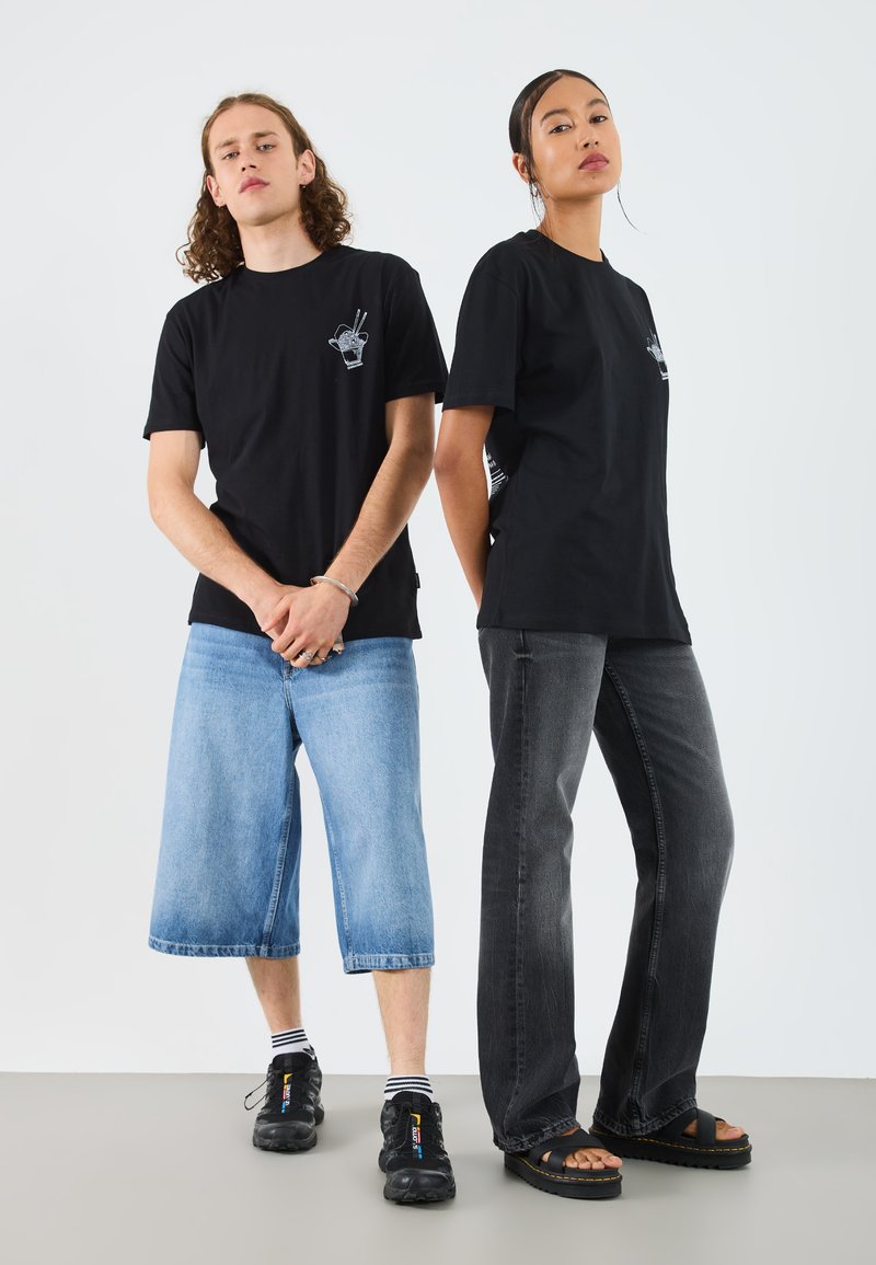 To personer iført svarte bomull t-skjorter med et håndgrafikk. Den ene har på en svart skjorte med grå jeans, den andre med blå vide shorts.