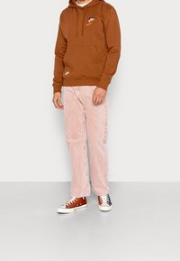 Sudadera con capucha marrón y bolsillo frontal, con logos bordados, combinada con pantalones de pana rosa claro y zapatillas bicolor.