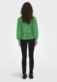 Femme aux longs cheveux bouclés portant un chemisier vert en dentelle, un jean skinny noir et des chaussures noires épaisses, debout de dos sur un fond blanc.