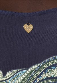 Le tissu côtelé bleu marine présente un motif coloré avec des accents verts et bleus. Une étiquette en forme de cœur doré affiche le nom de la marque.