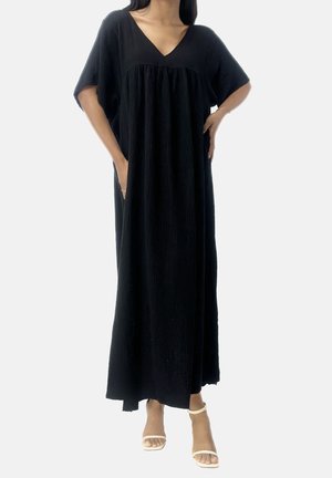 Mujer con un vestido negro largo y suelto con mangas cortas y escote en V, combinado con sandalias blancas con tira en el tobillo, de pie con las manos en los bolsillos.
