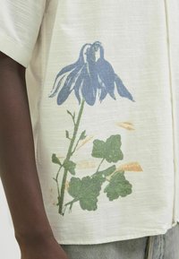 Short-sleeve, lightweight shirt in off-white with a blue floral design and green leaves. Features a subtle texture and relaxed fit.
