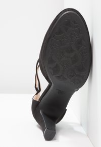 Chaussure à talon haut en suède noir avec un bout pointu et une bride au niveau de la cheville. La semelle présente un motif floral détaillé. Hauteur de talon approximative de 7 cm.