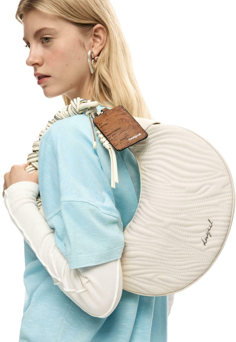 Mujer joven con cabello rubio que lleva mangas en capas de color azul claro y blanco, y sostiene un bolso blanco acolchado redondo con una etiqueta marrón.