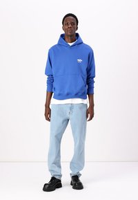 Blauer Hoodie mit Vordertasche, weißem Logo-Detail, kombiniert mit hellblauen Jeans und schwarzen, klobigen Schuhen vor einem schlichten Hintergrund.