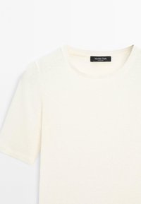 Krémový pletený top s krátkým rukávem a kulatým výstřihem, s černou značkou Massimo Dutti uvnitř límečku.