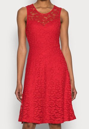 Ärmelloses rotes Spitzenkleid mit floralem Muster, figurbetonter Taille und rundem Ausschnitt, getragen von einer stehenden Person vor schlichtem Hintergrund.